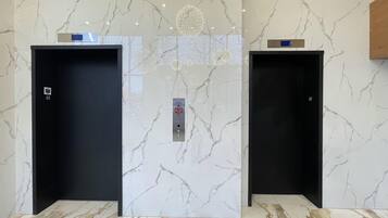 Two elevator doors with a marble wall and floor, a digital display above each door, and a control panel in the center.
