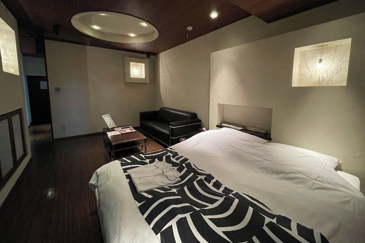 A modern hotel room with a large bed, a black leather sofa, a small table, and a headboard with built-in lighting.