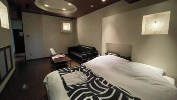 A modern hotel room with a large bed, a black leather sofa, a small table, and a headboard with built-in lighting.