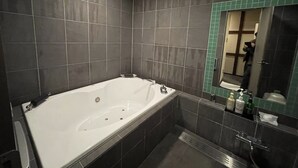 A modern bathroom with a large bathtub, dark tiled walls, and a small mirror with a green frame. - HOTEL Fuuga Luxury Resort - Adults Only (Maizuru)