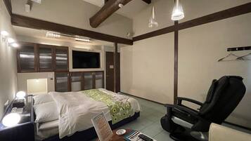 A hotel room with a bed, a desk with a lamp, a chair, a TV mounted on the wall, and a wooden ceiling beam.