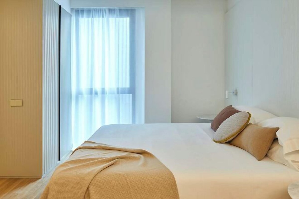 A modern hotel room with a large bed, a window with sheer curtains, and a neutral color scheme.