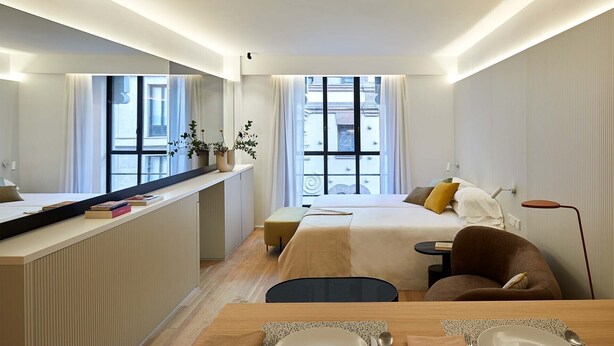 A modern hotel room with a bed, bedside table, chair, and a dining area with a table set for two. - 60 Balconies Iconic (Madrid)