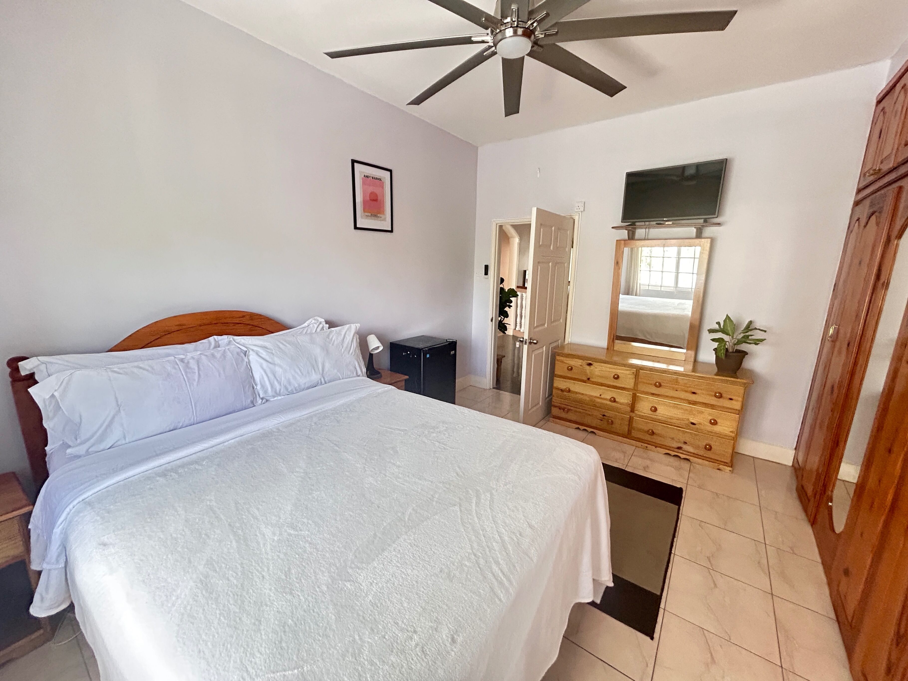 A bedroom with a bed, a ceiling fan, a dresser, a TV, and a door leading to another room.