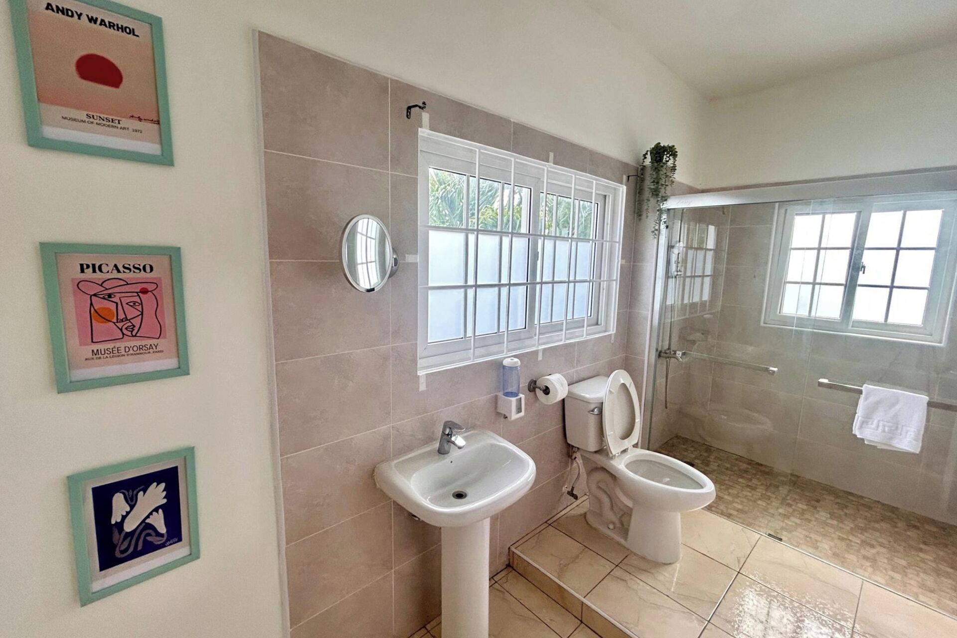 Une salle de bain avec une douche, deux lavabos et des toilettes. Des œuvres d’art encadrées ornent le mur.