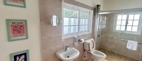 Une salle de bain avec une douche, deux lavabos et des toilettes. Des œuvres d’art encadrées ornent le mur.