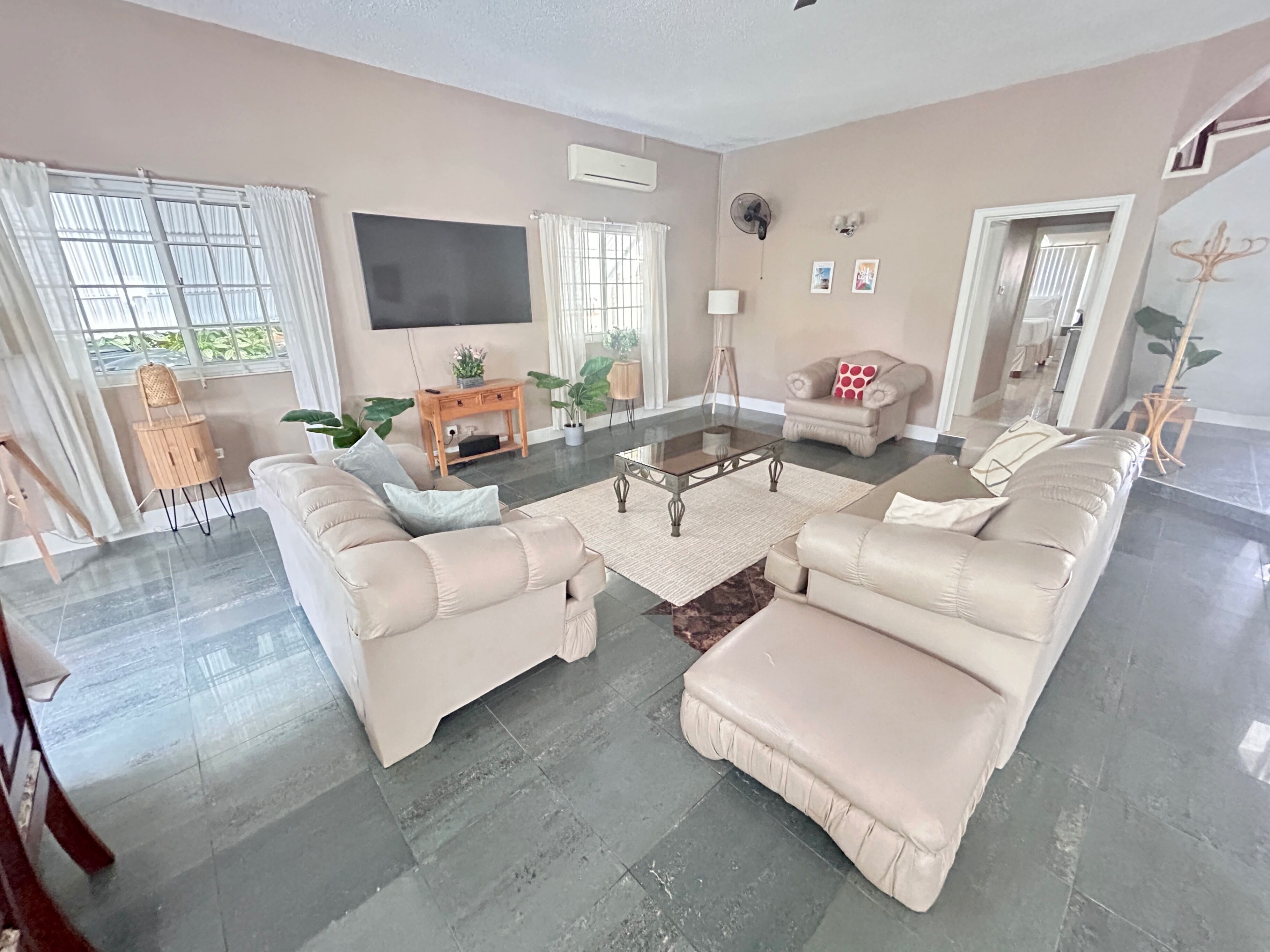 A spacious living room with a large sofa set, a coffee table, and a flat-screen TV mounted on the wall. There are potted plants and a small wooden side table.