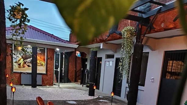 A courtyard with a wooden chair and table, a hanging plant, and a building with a red roof. - Hotel La Vid (La Fortuna)