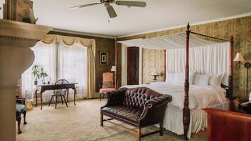 A four-poster bed with a canopy, a leather armchair, a wooden desk with a chair, and a ceiling fan in a room with floral wallpaper and a carpeted floor.