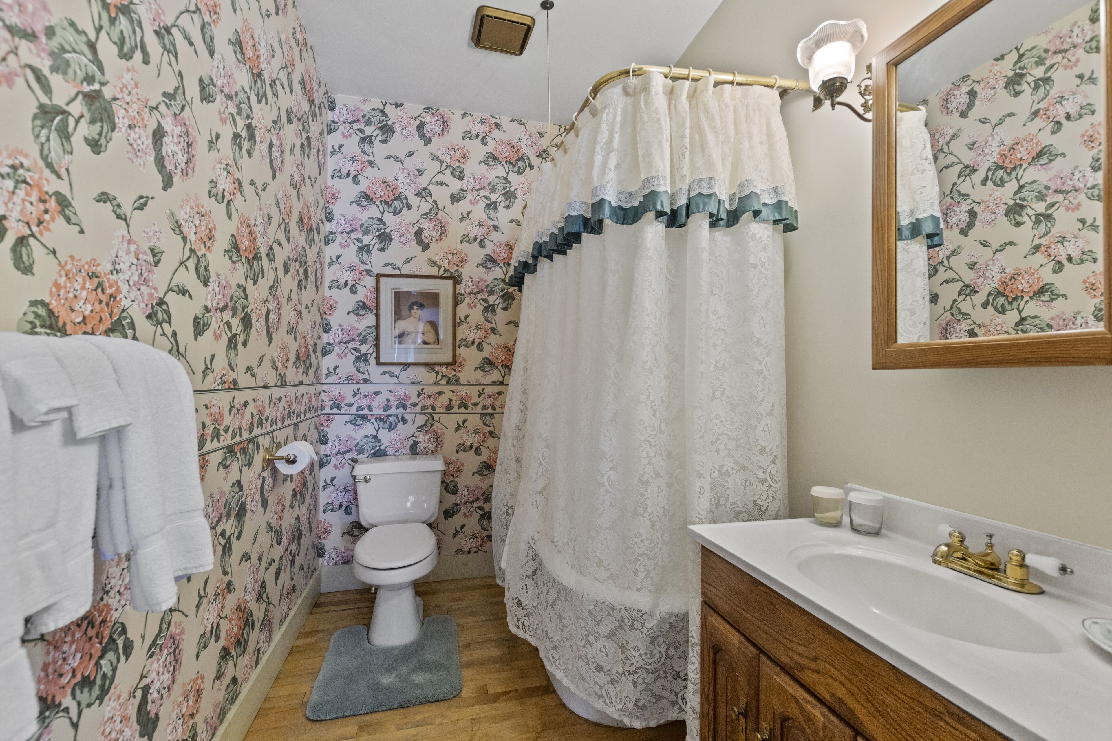 A bathroom with floral wallpaper, a white toilet, a shower curtain, a sink with a gold faucet, and a wooden cabinet.