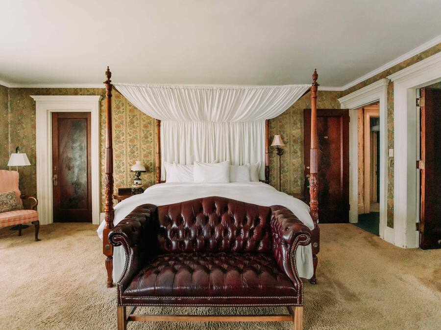A four-poster bed with a canopy, a leather armchair, and a classic chair in a room with a carpeted floor and floral wallpaper.