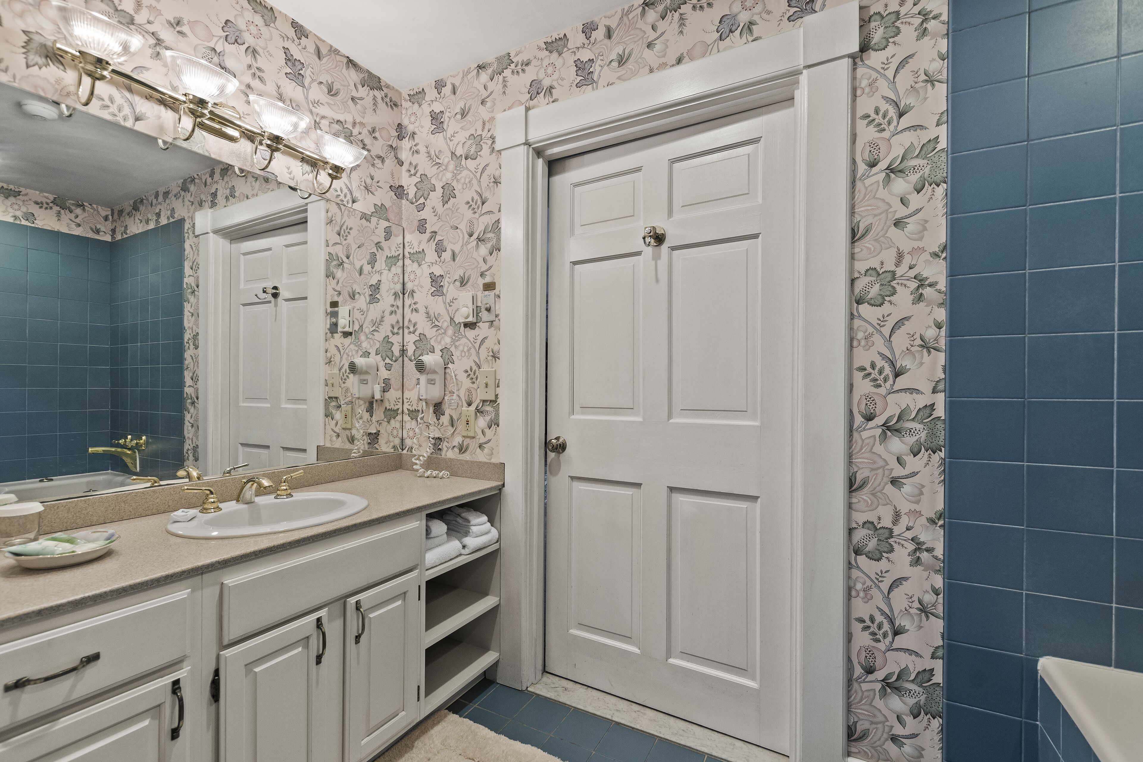 A bathroom with a white vanity, a sink, and a mirror. There is a door with a handle and a towel rack above it. The walls have a floral wallpaper pattern.