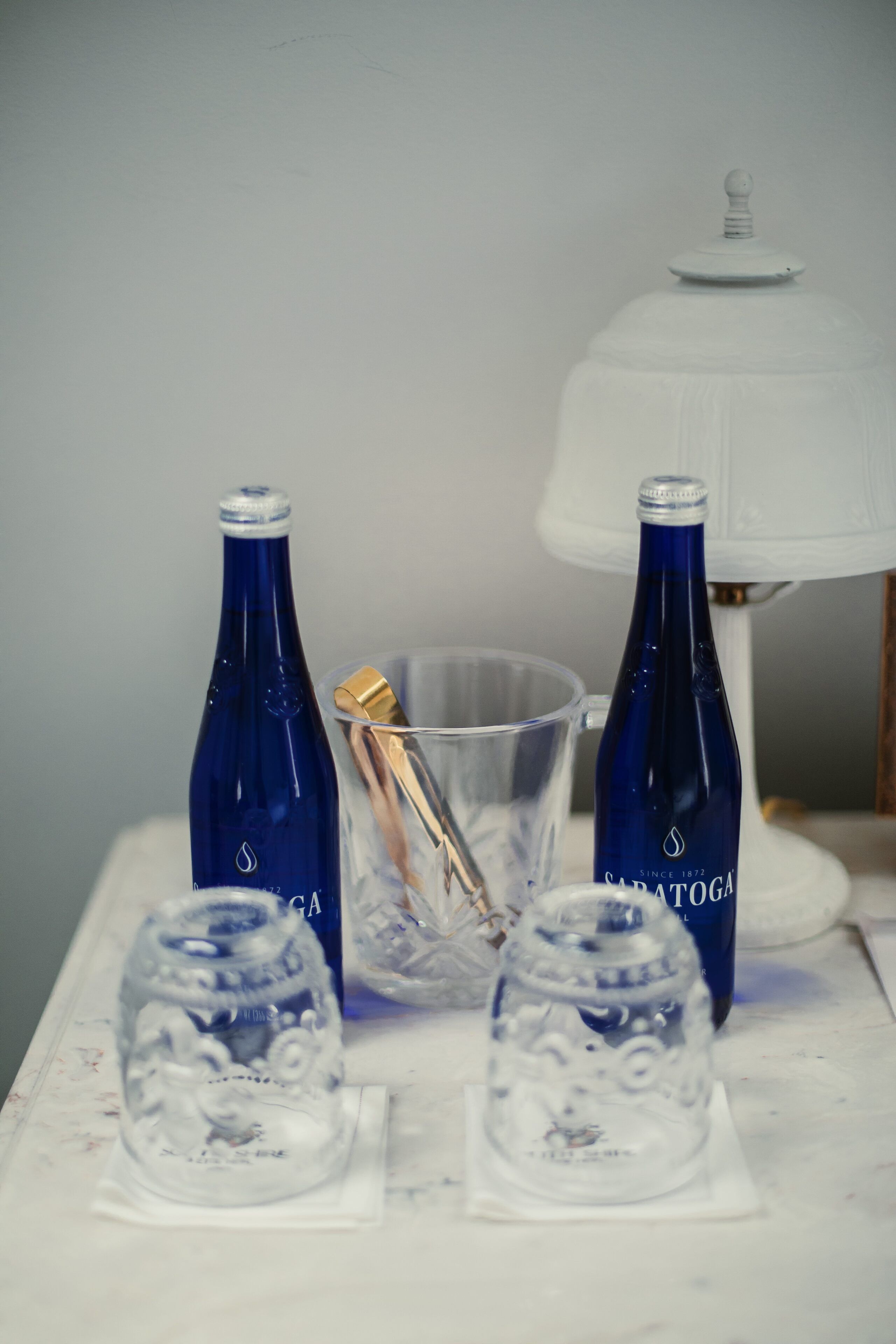Una mesa con dos botellas azules, un vaso transparente y un vaso decorativo sobre una superficie blanca.