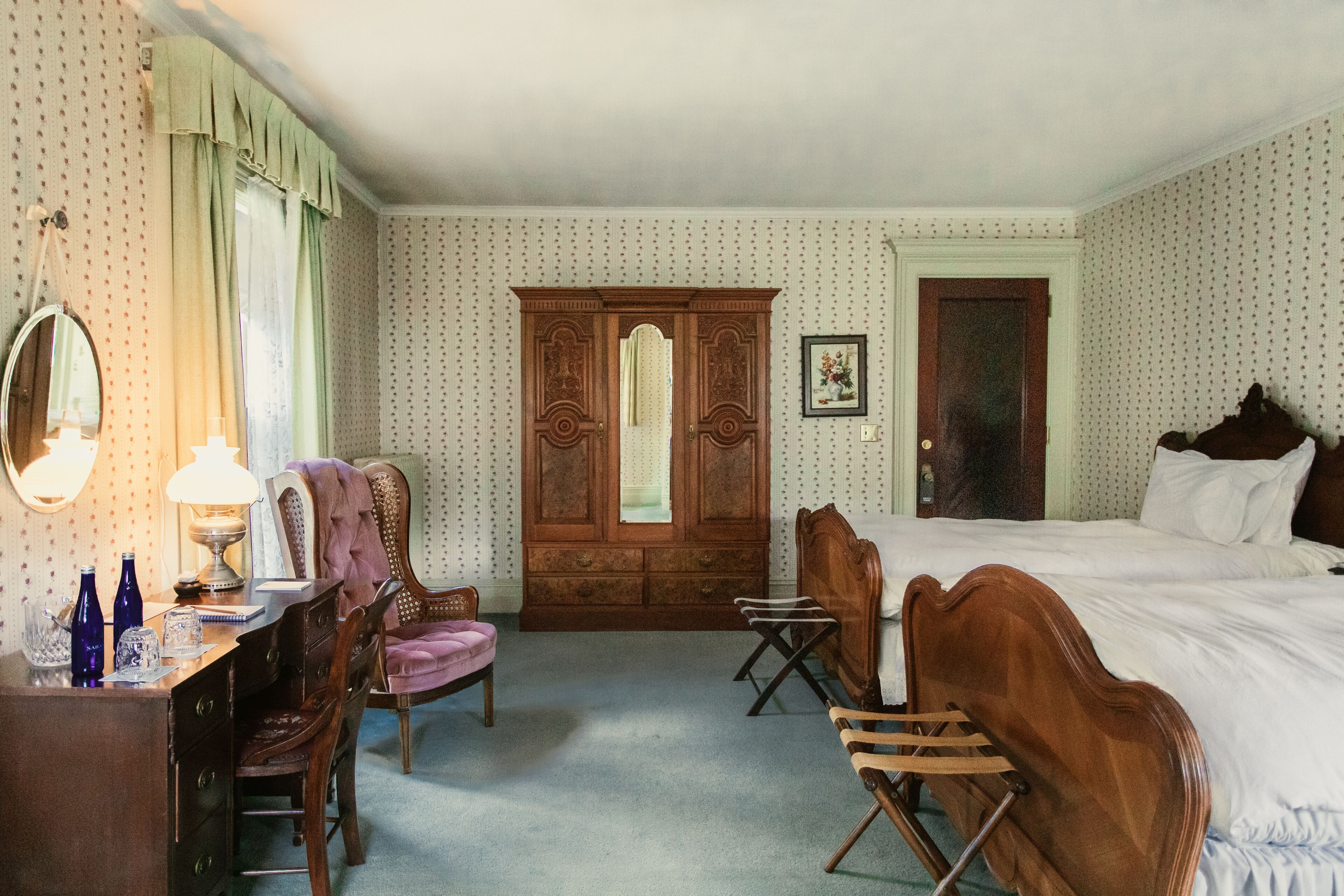 A hotel room with a large wooden wardrobe, a desk with a lamp, a chair, and a bed.