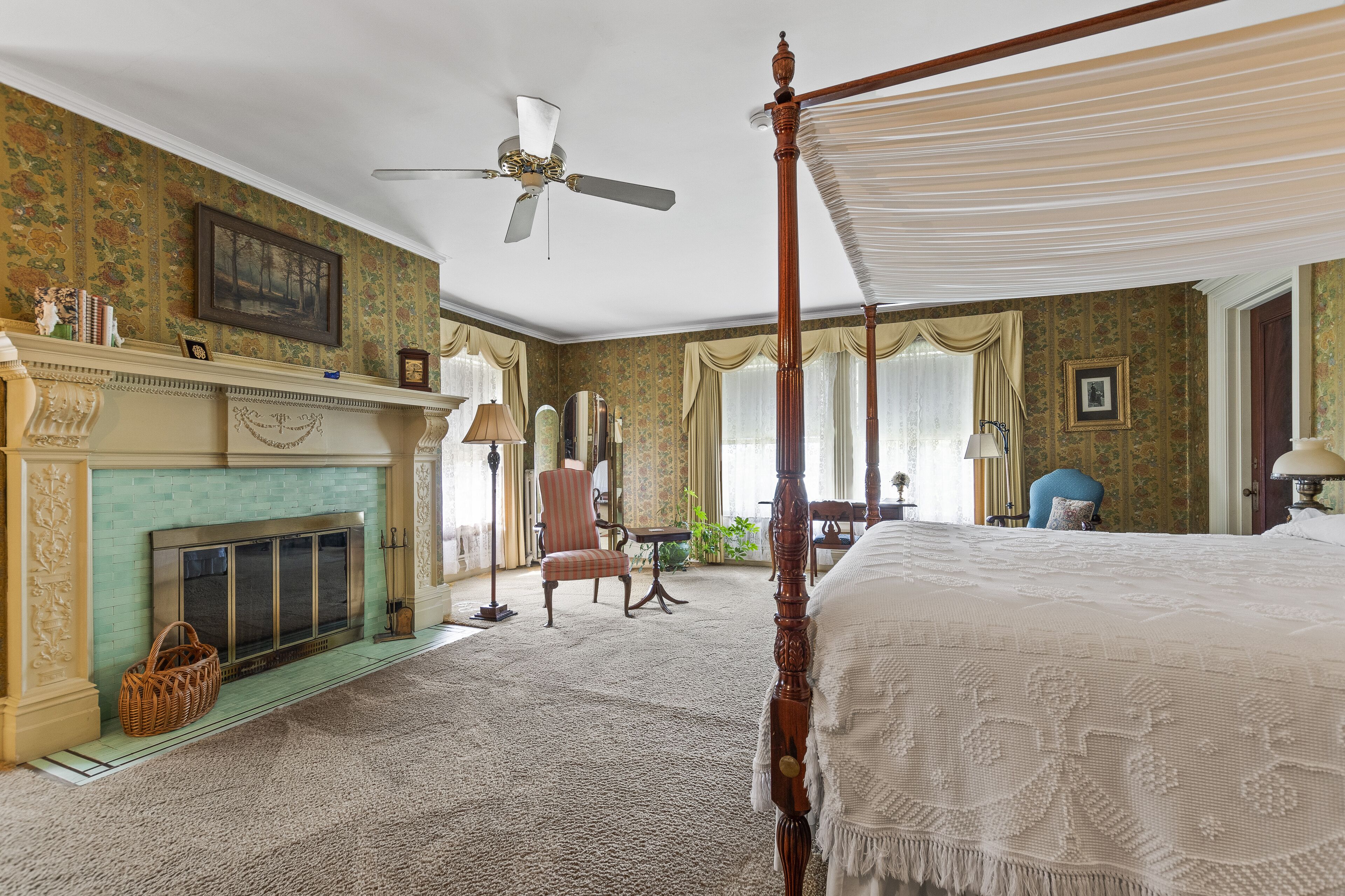 A spacious bedroom with a four-poster bed, a fireplace, a ceiling fan, and a large window with curtains.