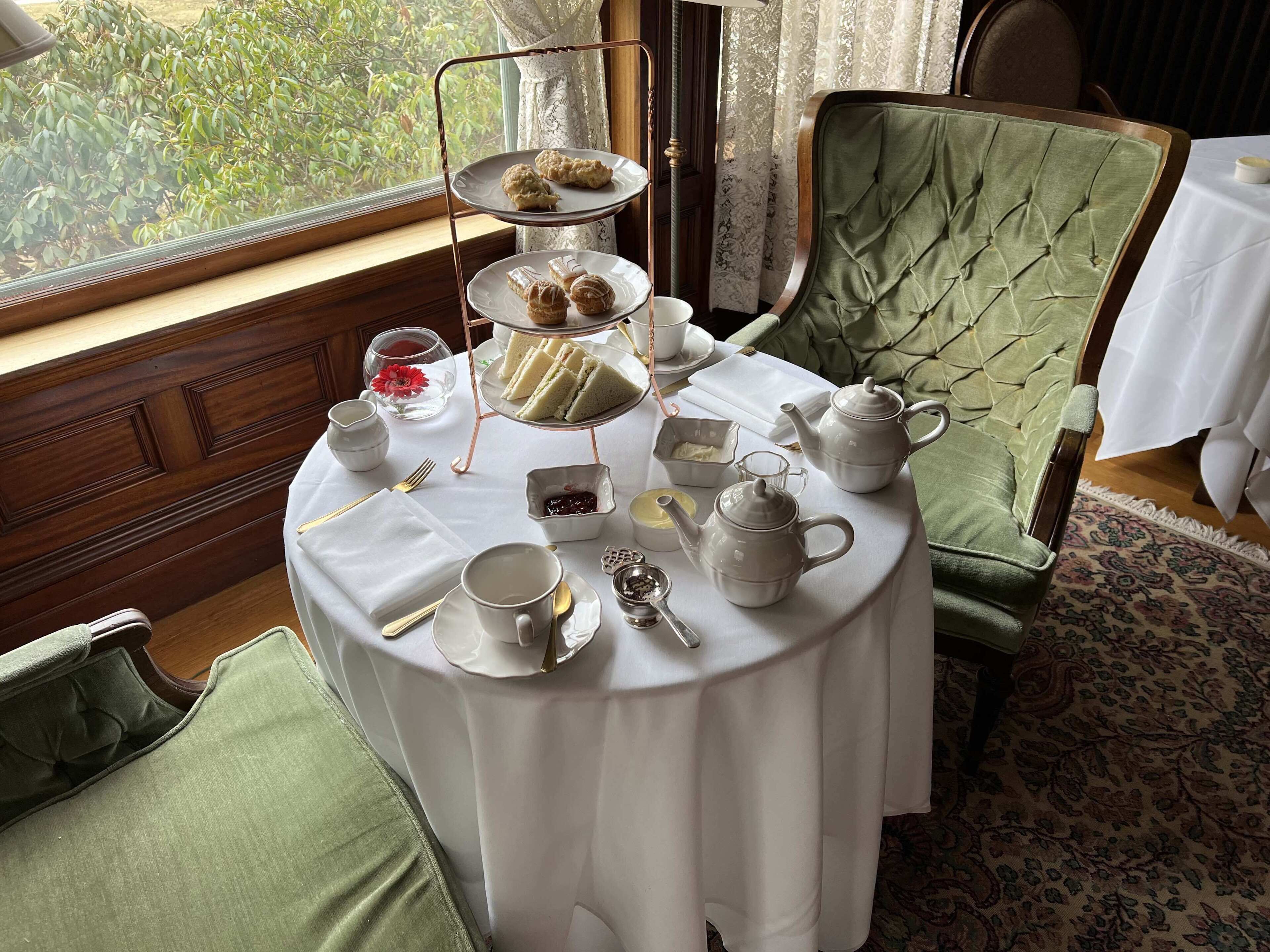 Une table de salle à manger ronde, agrémentée d’un présentoir à gâteaux à plusieurs niveaux, d’un service à thé et d’une chaise recouverte d’un tissu vert.
