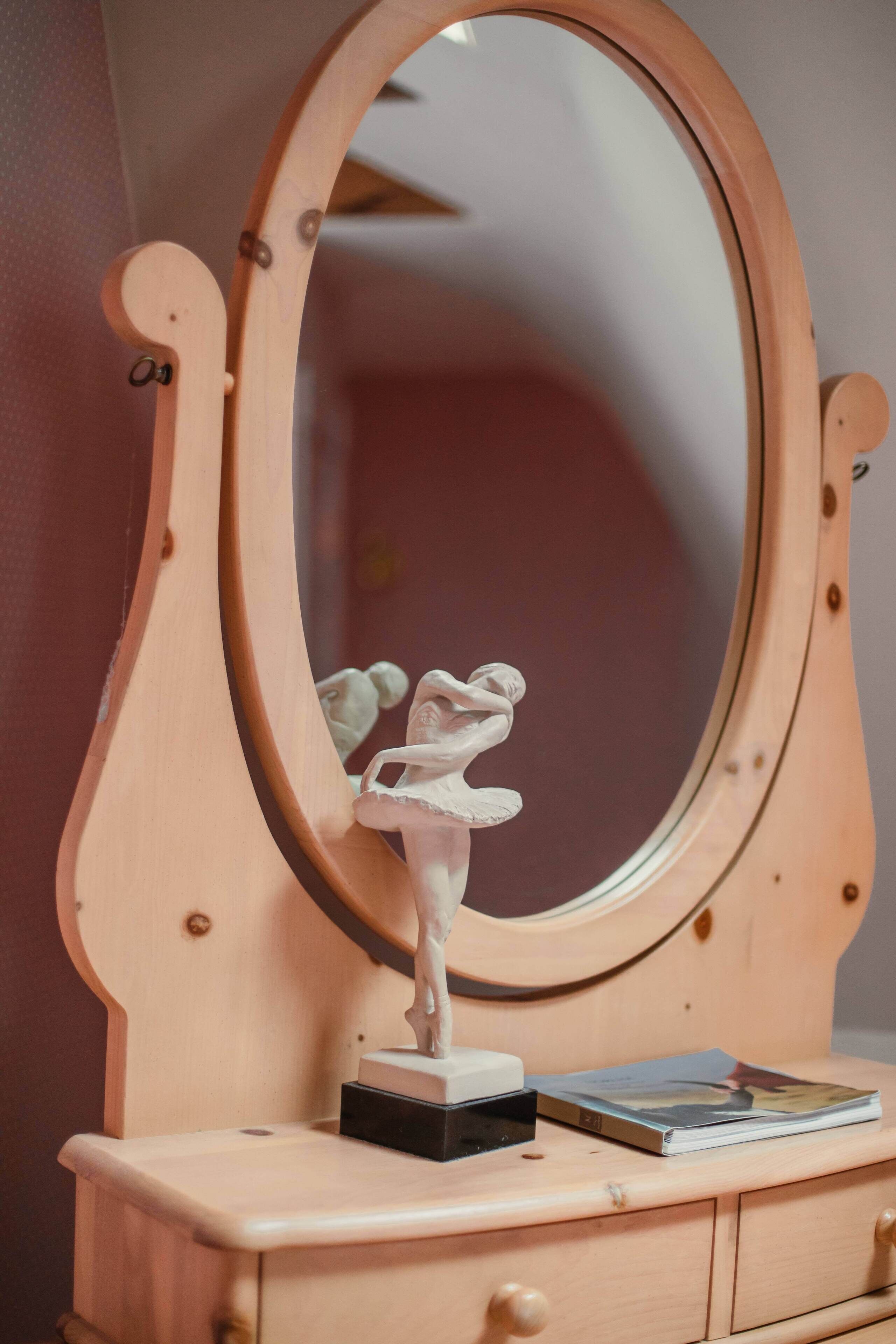 A wooden dresser with a mirror, a ballet dancer figurine, and a book.
