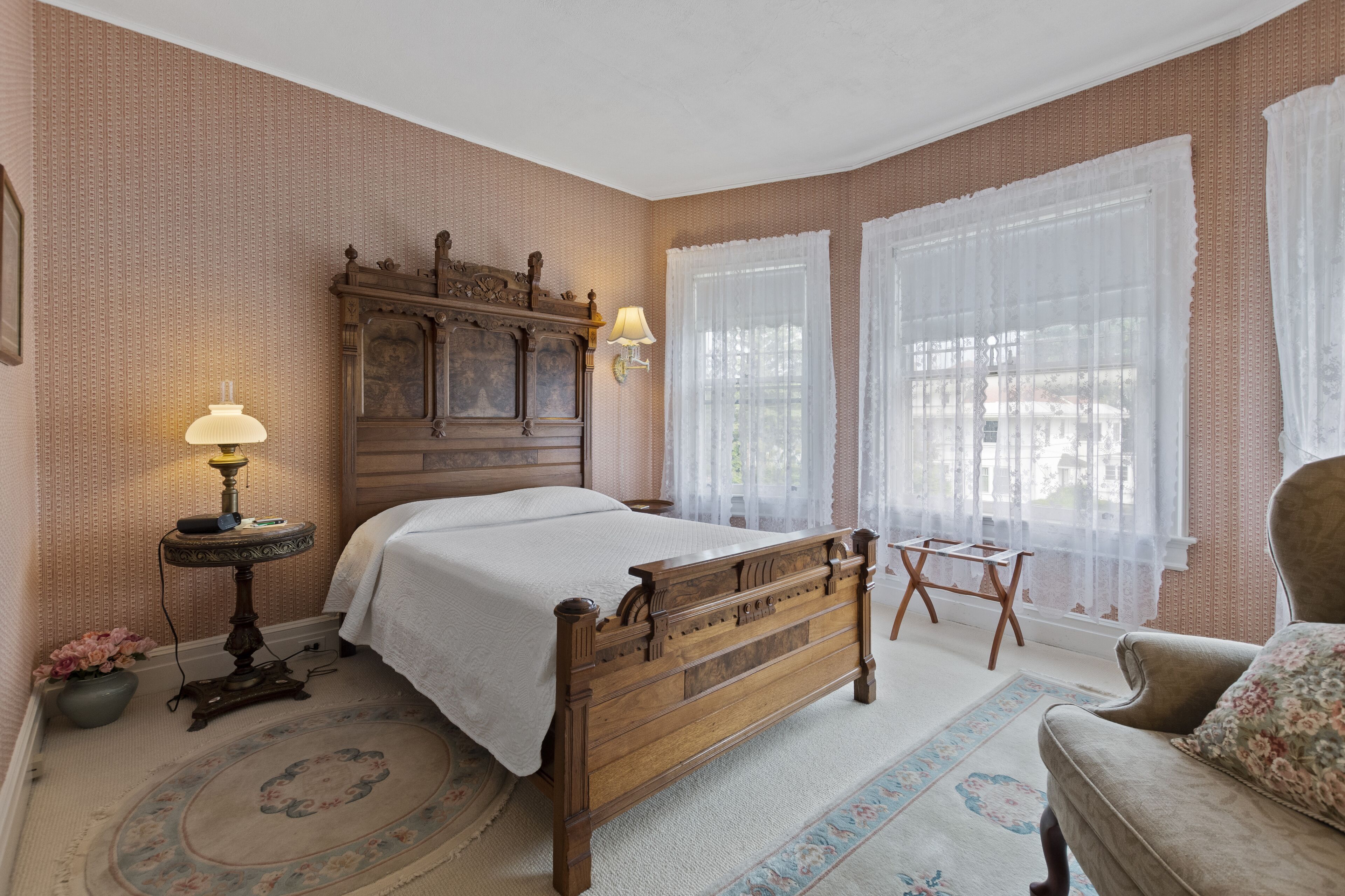 A bedroom with a wooden bed, a bedside table with a lamp, a chair, and a window with sheer curtains.