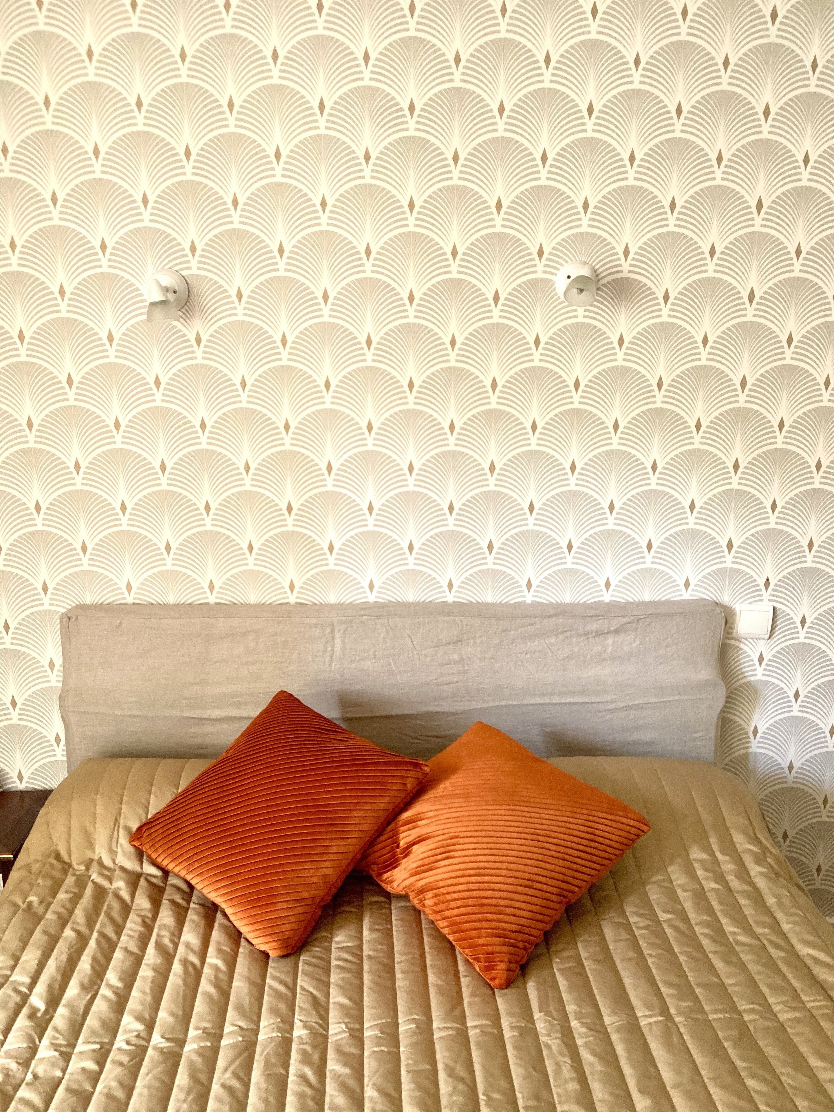 A bed with a beige quilt and two orange pillows, set against a wall with a repeating geometric pattern.