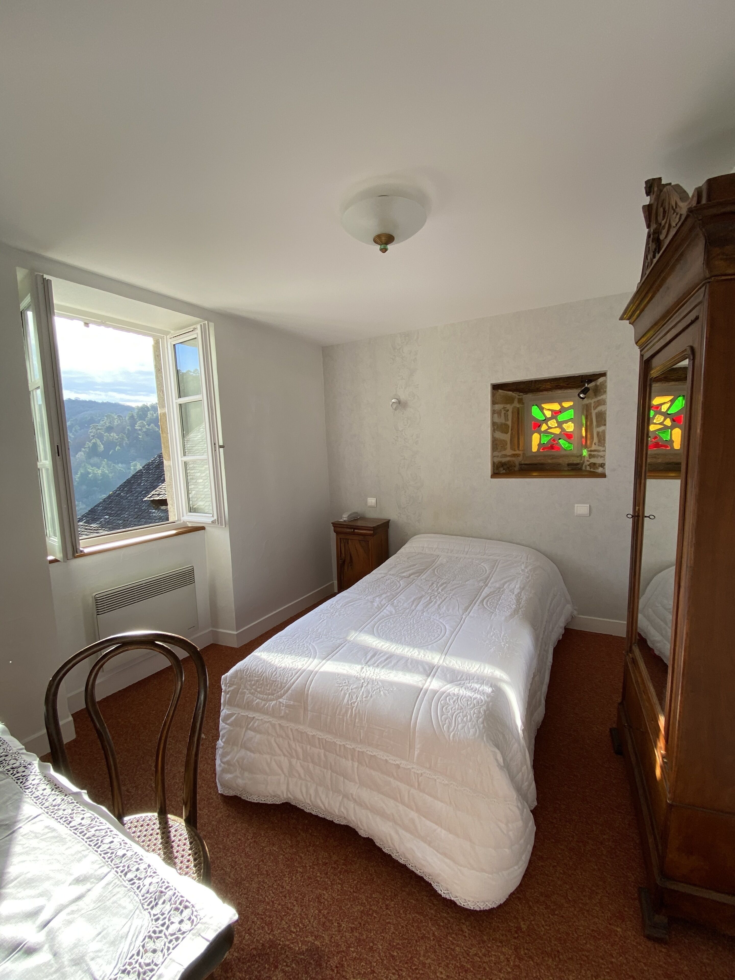 A bedroom with a large bed, a wooden chair, a dresser, and a window with a view of a forested area.