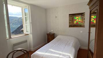 A bedroom with a large bed, a wooden chair, a dresser, and a window with a view of a forested area.