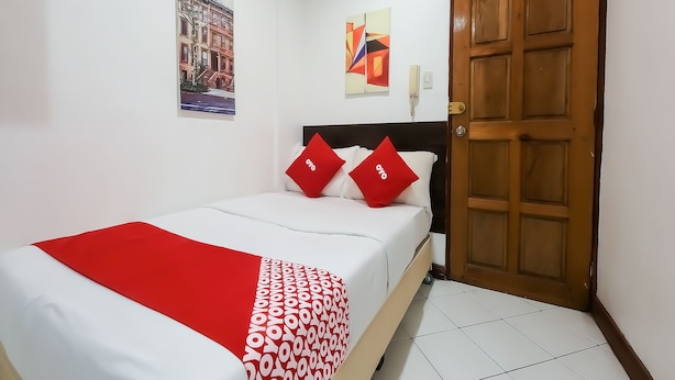 Una cama bien hecha con una colcha de cuadros rojos y blancos, dos almohadas rojas con la palabra "oyo" y una puerta de madera a la derecha.