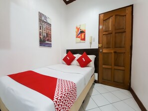 Cama bem arrumada com colcha estampada em vermelho e branco, duas almofadas vermelhas com a palavra "oyo" e uma porta de madeira à direita.