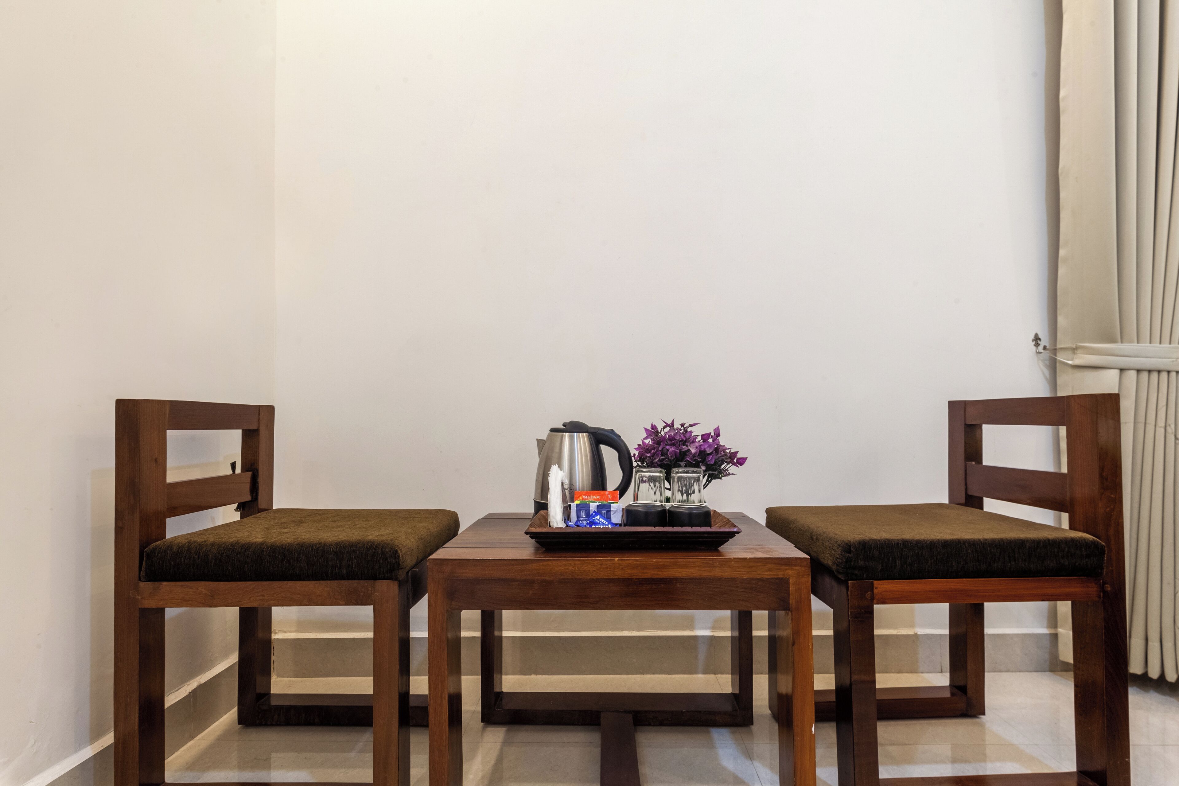 A small, neatly arranged room with two wooden chairs and a table, featuring a kettle, a tray with a bottle and glasses, and a vase with flowers.