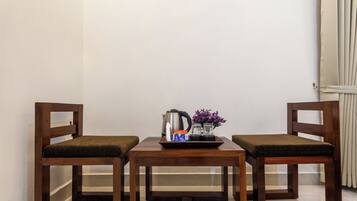 A small, neatly arranged room with two wooden chairs and a table, featuring a kettle, a tray with a bottle and glasses, and a vase with flowers.