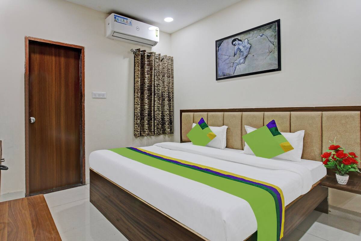 A hotel room with a wooden door, air conditioning unit, patterned curtains, a bed with a green and purple bedspread, a headboard with decorative pillows, a framed artwork on the wall, and a small table with a vase of red flowers.