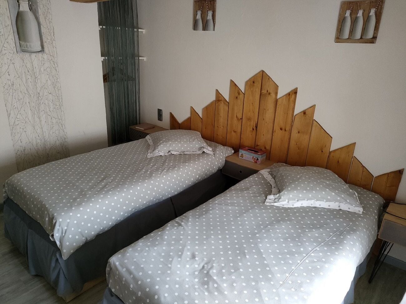 A room with two single beds, wooden headboards, a small bedside table with a lamp, and a wall-mounted shelf with bottles.