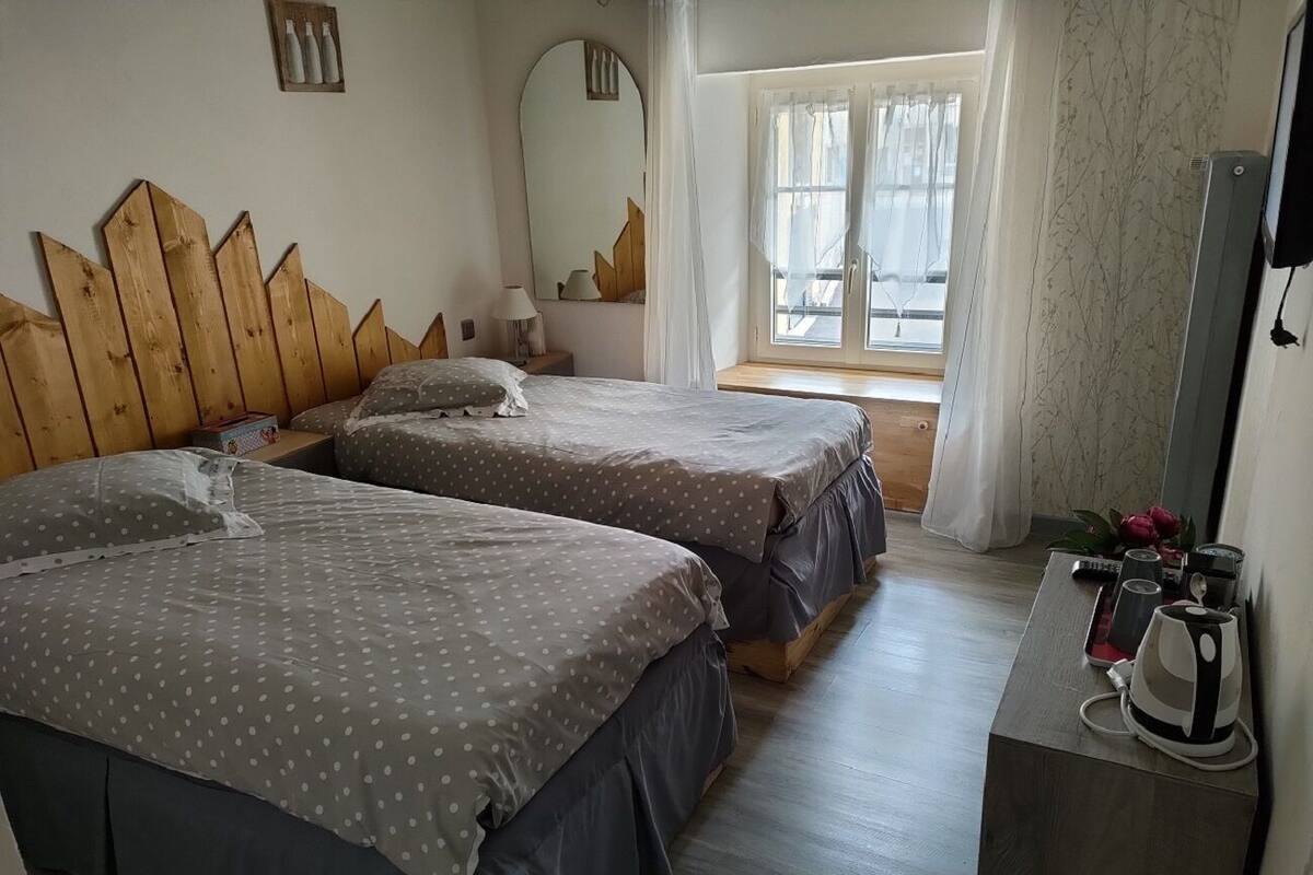 Ein Schlafzimmer mit zwei Betten, einem hölzernen Kopfteil, einem Nachttisch mit Wasserkocher und einem Fenster mit durchsichtigen Vorhängen.