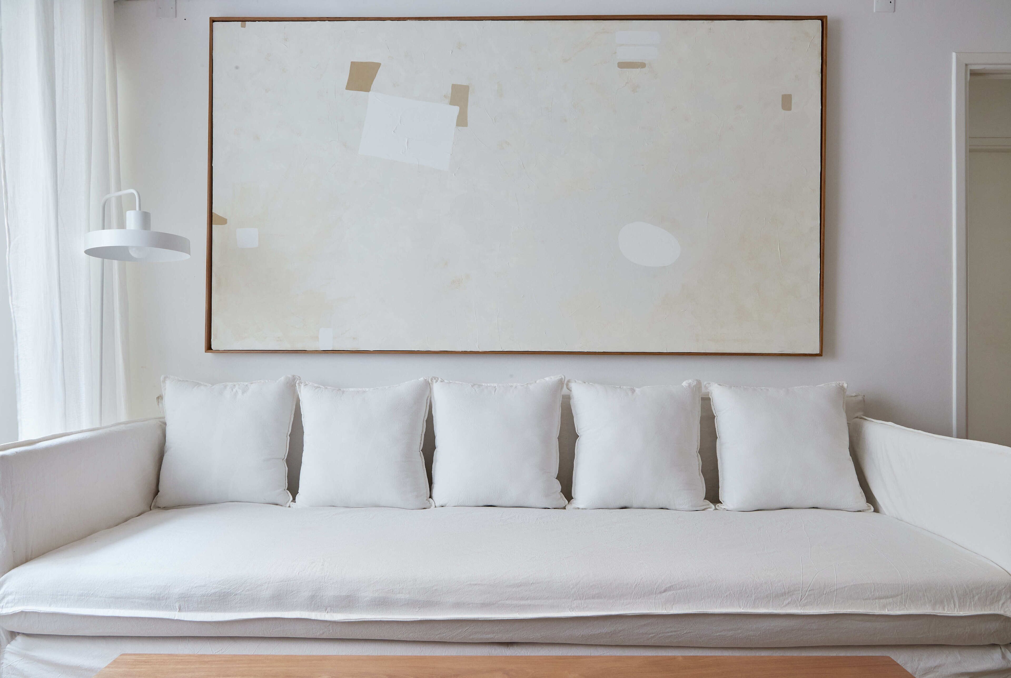 A white sofa with five cushions, a white wall, and a large abstract painting.