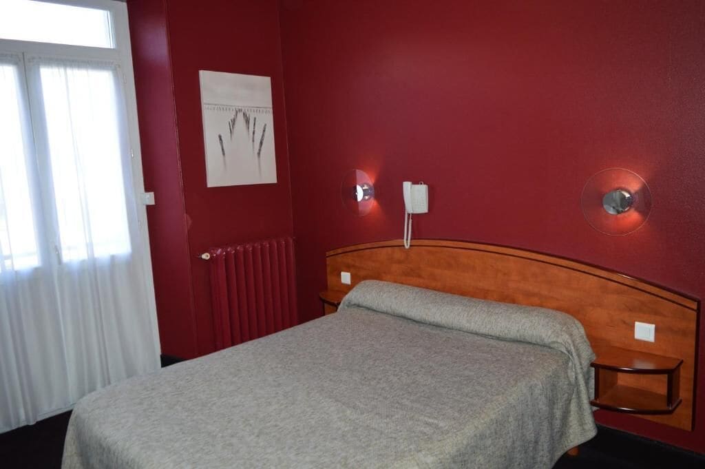 A hotel room with a single bed, a radiator, a wall-mounted light, and a window with curtains.