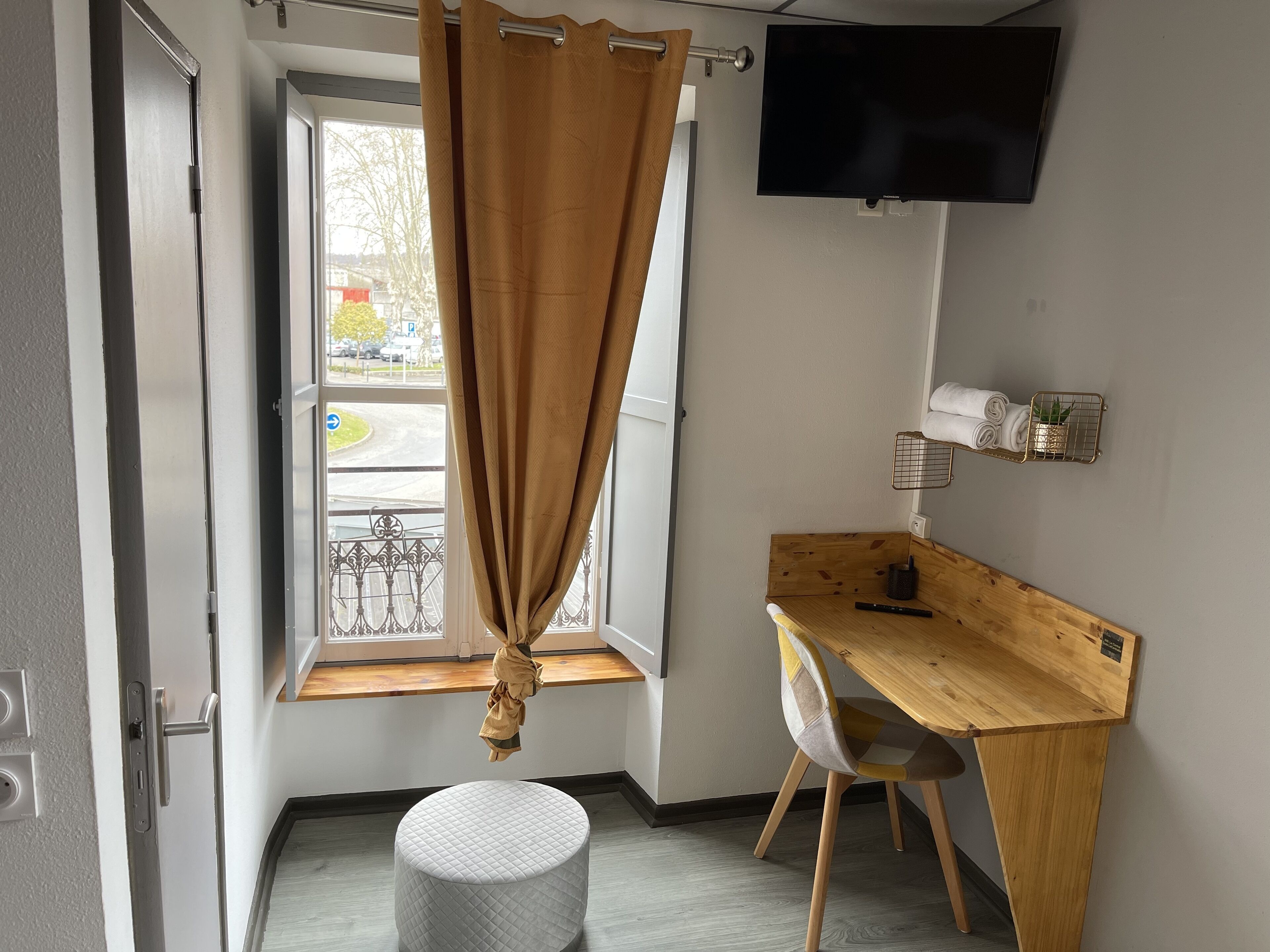 A room with a wooden desk, a chair, a round ottoman, a TV mounted on the wall, and a window with a curtain.