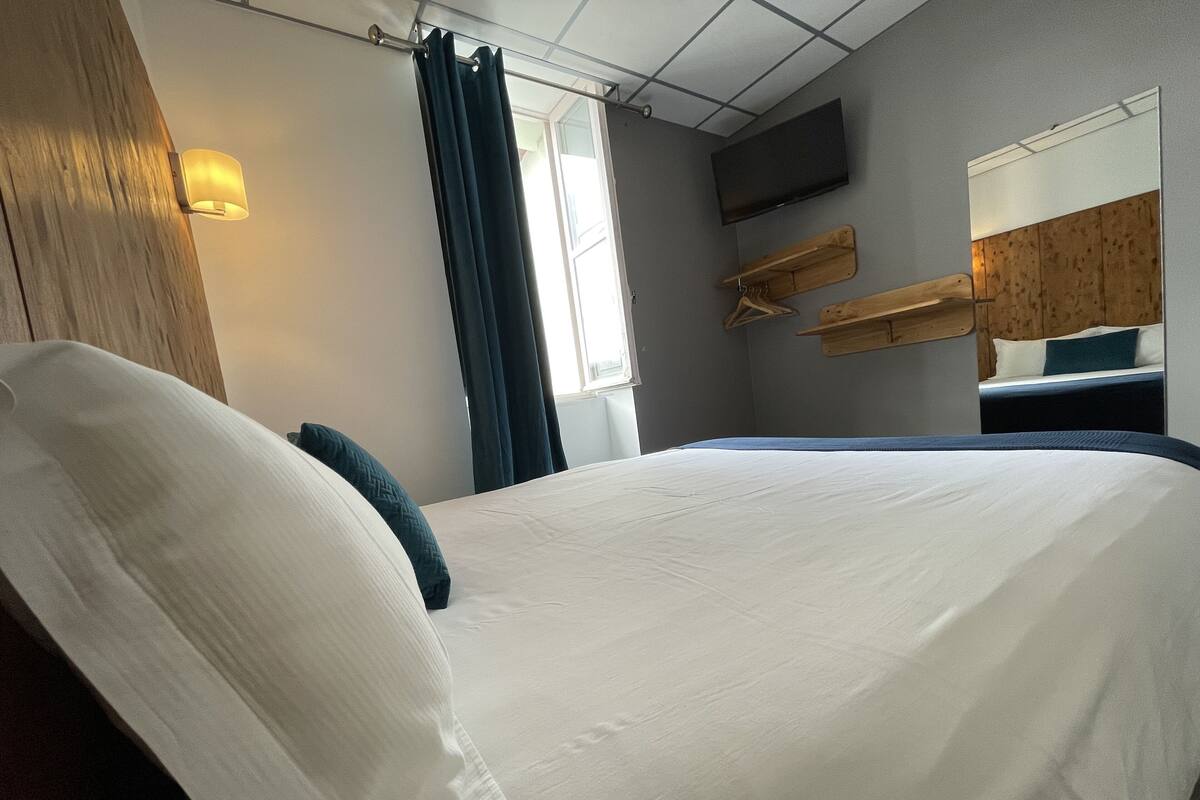 Une chambre d’hôtel avec un lit, une tête de lit en bois, un téléviseur fixé au mur et une fenêtre avec des rideaux.