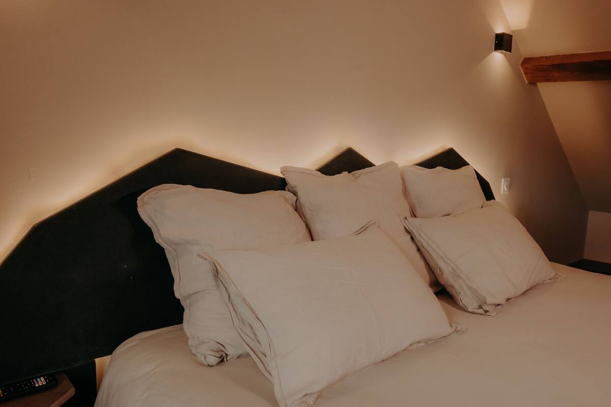 Un lit bien fait, avec du linge de lit blanc et des oreillers, une tête de lit sombre et un luminaire fixé au mur.
