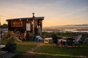 A rustic cabin with a wooden exterior, a small deck, outdoor seating, and a view of the beach and ocean at sunset.