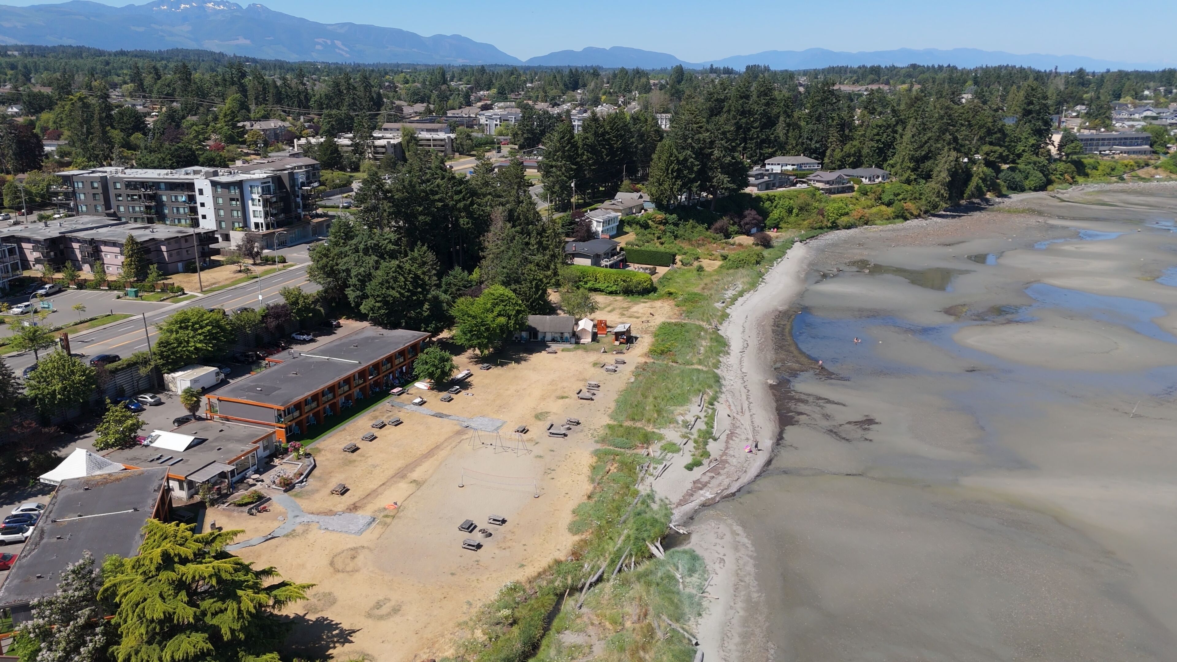An aerial view of a coastal area with a sandy beach, buildings, and a forested hillside.