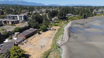 An aerial view of a coastal area with a sandy beach, buildings, and a forested hillside.