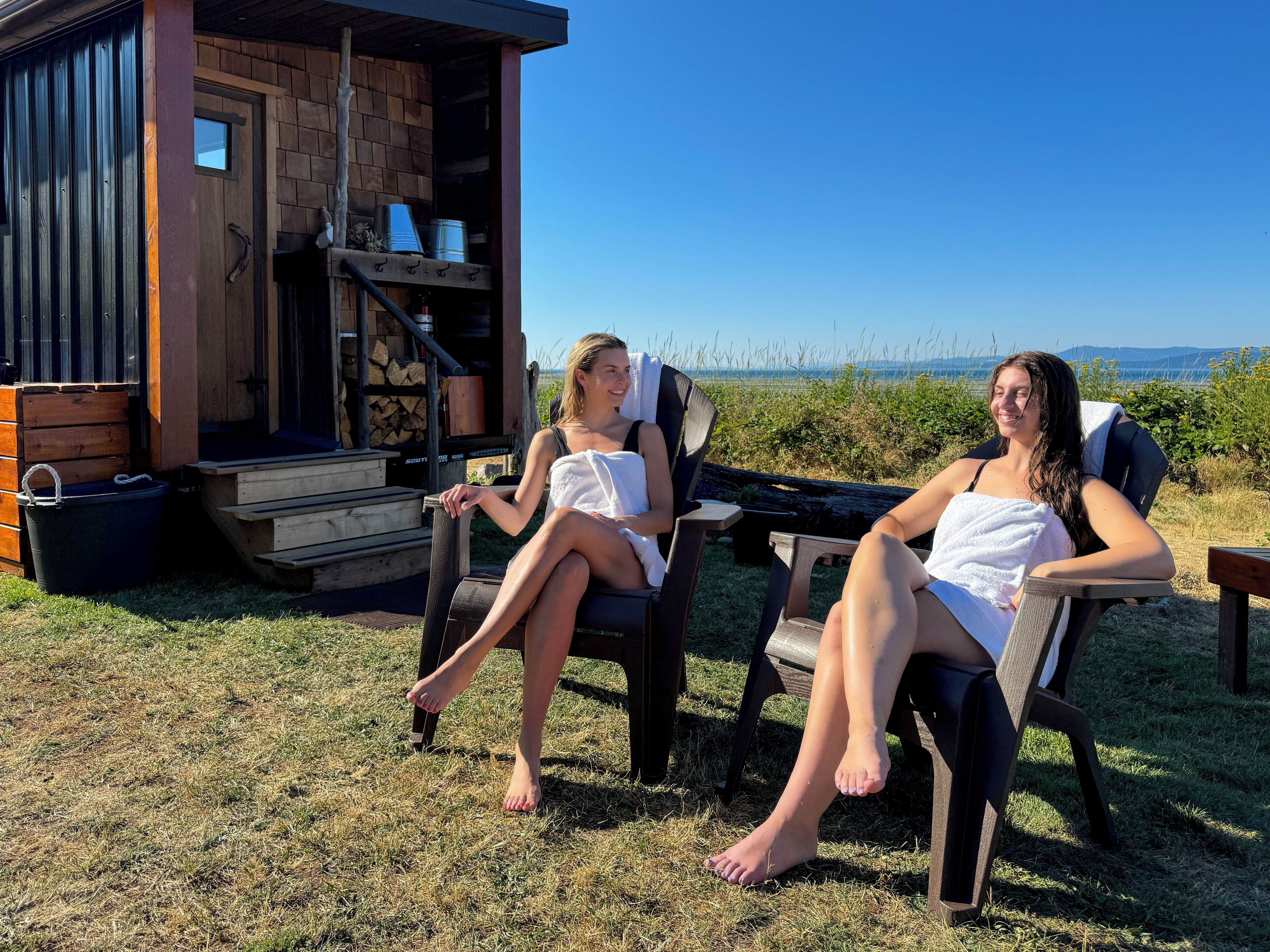 Zwei Personen sitzen auf Stühlen vor einer Holzhütte mit freiem Blick auf die Landschaft.
