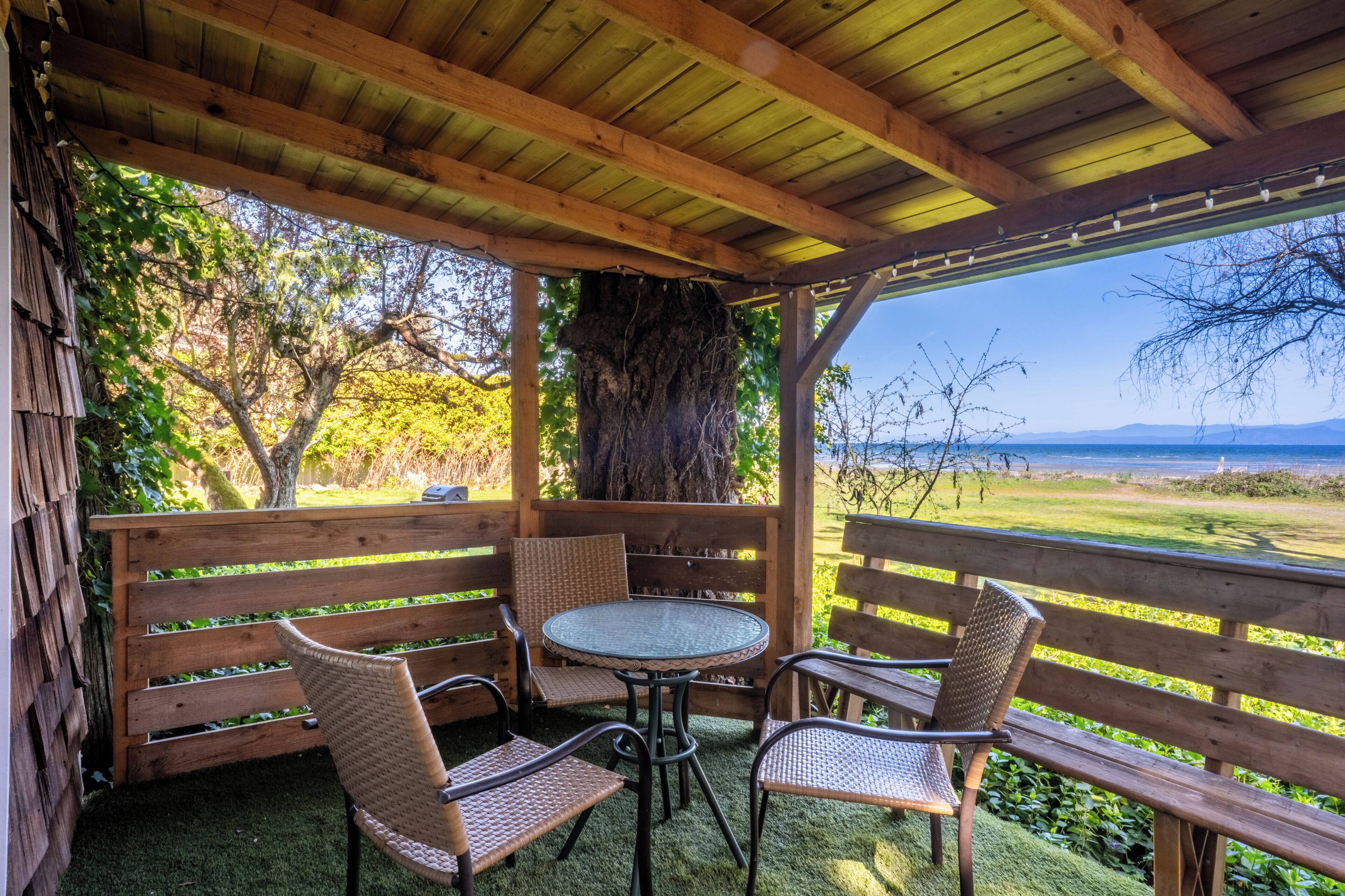 A covered patio with a round table and wicker chairs, surrounded by wooden railings and a view of a grassy landscape.