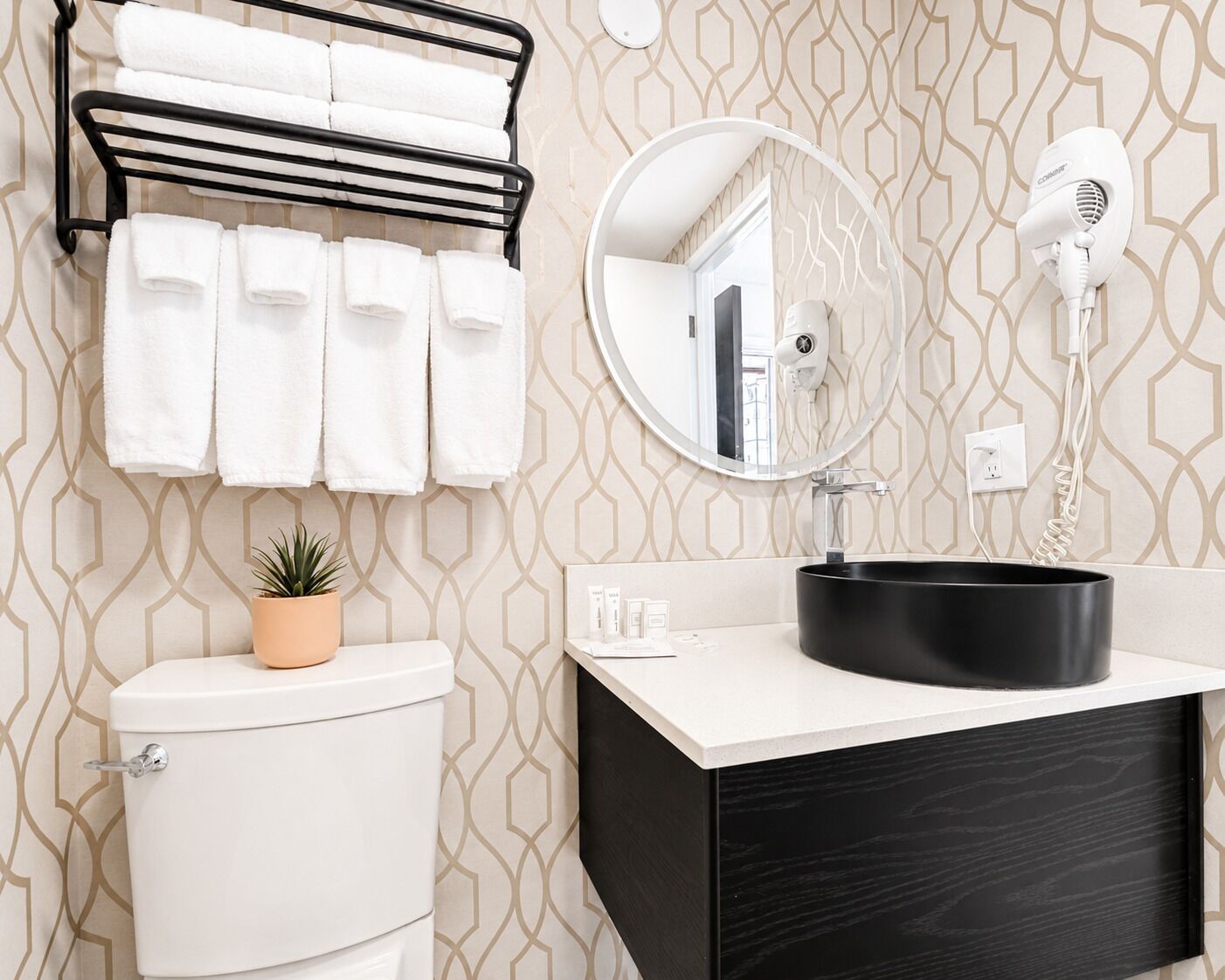 Une salle de bain comprenant des toilettes, un lavabo avec un receveur noir, un porte-serviettes avec des serviettes blanches, une plante en pot et un sèche-cheveux fixé au mur.