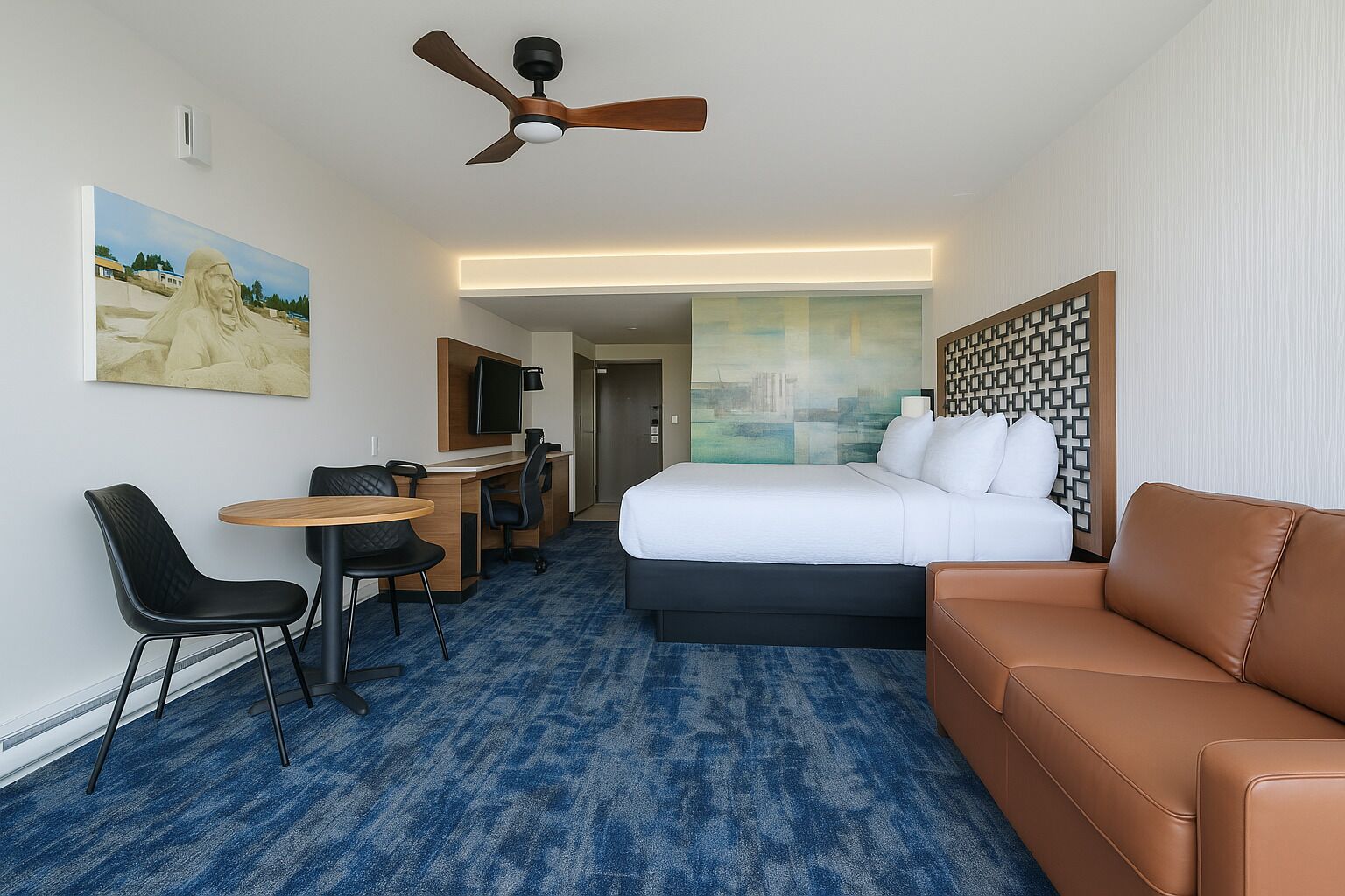 A hotel room with a bed, a desk with a chair, a sofa, a ceiling fan, and a painting on the wall.