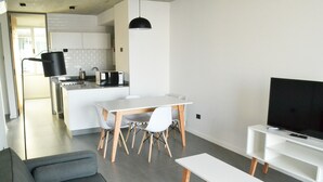 Ein modernes Wohnzimmer mit grauem Sofa, einem weißen Esstisch mit Stühlen, einem Flachbildfernseher auf einem weißen Schrank und einer Küche im Hintergrund.