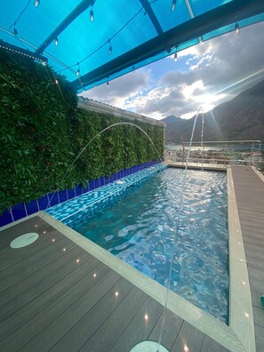A swimming pool with a wooden deck, surrounded by a blue-tiled edge and a green wall with plants. - Hotel Palmera Dorada (Santiago Atitlán)