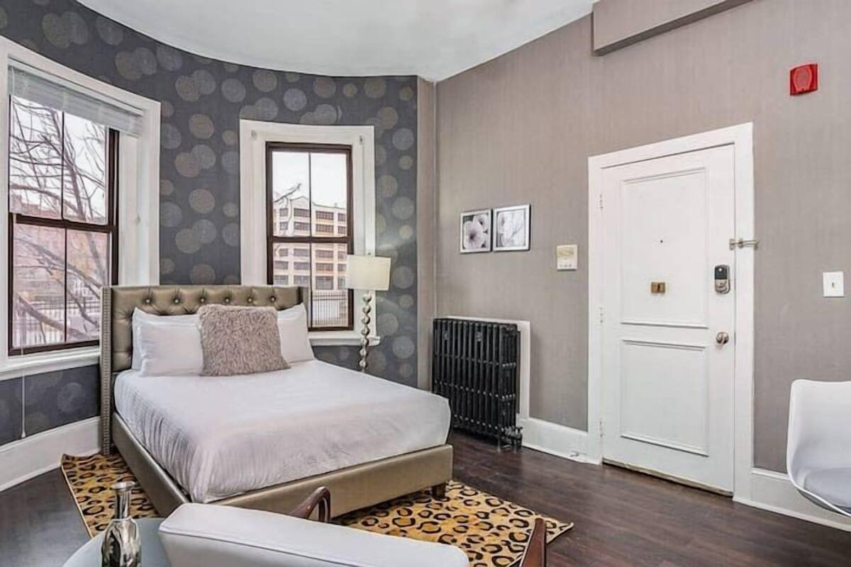 A bedroom with a large bed, a radiator, a white door, and a window with a view of buildings.