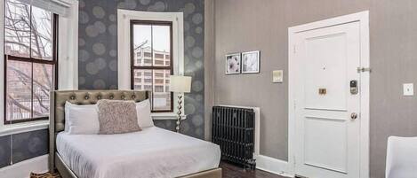 A bedroom with a large bed, a radiator, a white door, and a window with a view of buildings.