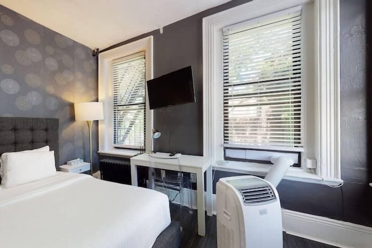 A hotel room with a large bed, a desk, a chair, a TV, and two windows with blinds.