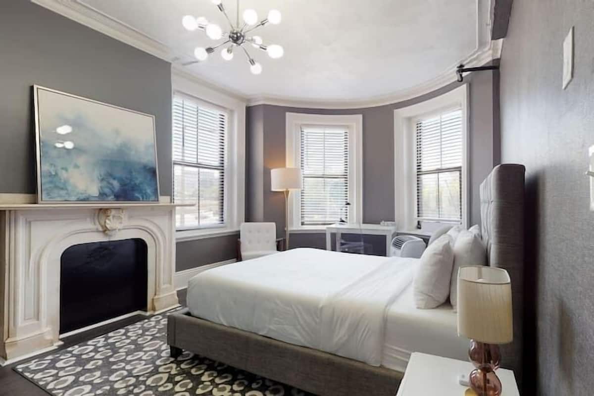 A modern bedroom with a large bed, a fireplace, and two windows with blinds.